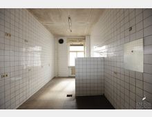 Ein leerer Raum mit gefliesten Wänden und gefliestem Boden, hellem Tageslicht durch ein einfaches Fenster. An der Decke befindet sich eine Leuchtstofflampe, und in der Wand ist ein kleiner Lüfter eingebaut.