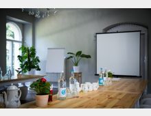 Heller Seminarraum mit einem großen Holztisch, darauf stehen Wasserflaschen, Gläser, Teekannen und Blumen in Töpfen. Im Hintergrund befinden sich ein Flipchart, eine Leinwand sowie Pflanzen und ein großes Fenster mit Tageslichteinfall.