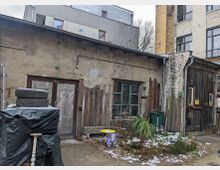 Hinterhof mit einem alten, verwitterten Gebäude aus Putz- und Holzelementen, teilweise mit Brettern verkleidet. Im Vordergrund liegen Reifen, eine Plane, ein kleiner gelber Kindertisch und etwas Schnee auf dem Boden, umgeben von Pflanzenresten und Mülltonnen.