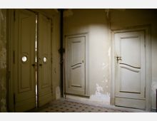 Ein Innenraum mit alten, beigen Holztüren; eine doppelflügelige Tür mit kleinen runden Fenstern links und zwei schmale Einzeltüren rechts. Die Wände sind abgenutzt und teilweise mit abgeblätterter Farbe, der Boden hat ein geometrisches Fliesenmuster.