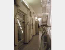 Innenansicht eines Gefängnisflurs mit cremefarben gestrichenen Wänden. Zu sehen sind Zellentüren mit kleinen Fenstern, Überwachungseinrichtungen und eine Metallbrüstung, die entlang eines Ganges führt.