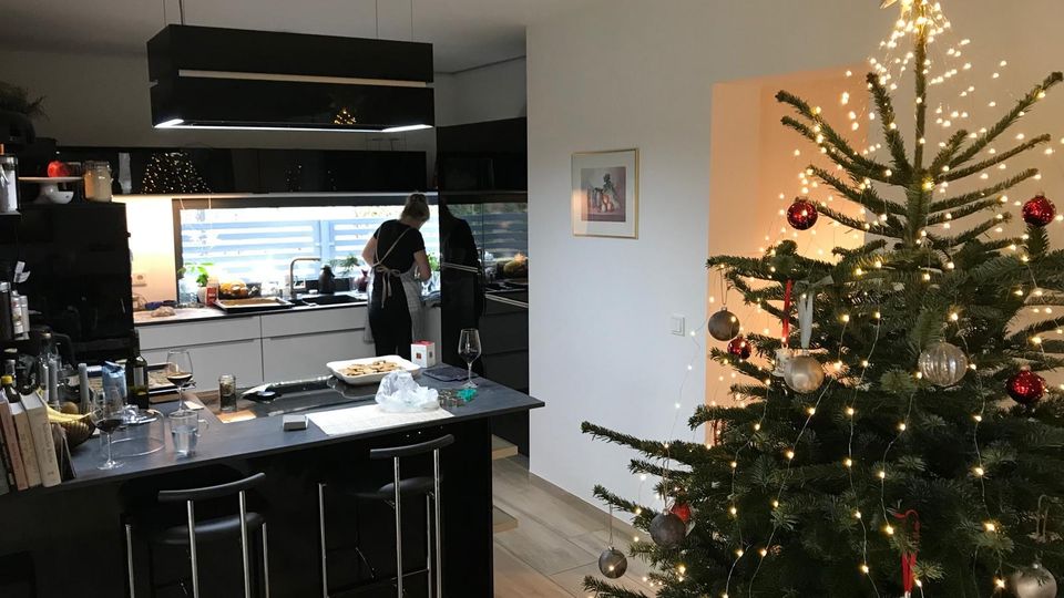 Moderne Wohnküche mit dunkler Kücheninsel und Barhockern, schwarzen Hängelampen sowie einer langen Fensterfront über der Arbeitsfläche. Rechts steht ein großer, mit Lichterkette, Kugeln und Sternspitze geschmückter Weihnachtsbaum vor einer weißen Wandnische.