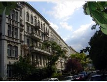 Straßenszene mit historischen, mehrstöckigen Wohnhäusern im Gründerzeitstil, die mit reich verzierten Fassaden und kleinen Balkonen gestaltet sind; im Vordergrund Bäume und parkende Autos, teils unter einer bewölkten, blauen Himmel.