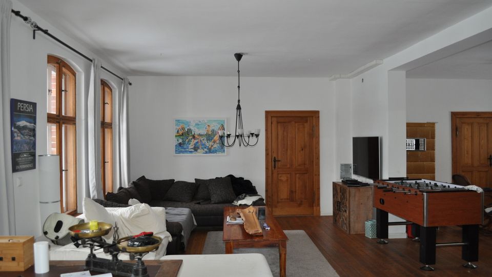 Wohnzimmer mit Holzboden, weißen Wänden und mehreren Holztüren. Der Raum enthält eine große dunkle Eckcouch, einen Holztisch, einen Tischkicker und eine Wanddekoration mit Gemälde, sowie einen Kronleuchter an der Decke. Drei große Fenster mit Holzrahmen lassen Licht herein.