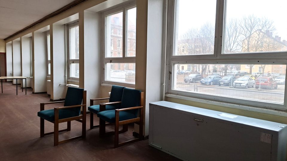 Langer Innenraum mit einer Reihe großer Fenster entlang der Wand, braunem Boden und spärlicher Möblierung mit zwei Sesseln und einem Tisch. Durch die Fenster sind eine Straße mit geparkten Autos sowie gegenüberliegende Gebäude und kahle Bäume zu sehen.