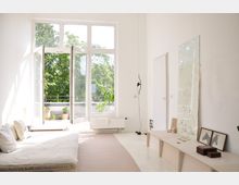 Helles Wohnzimmer mit großen, bodentiefen Fenstern, die auf einen Balkon mit Pflanzen führen. Ein schlichtes Sofa mit weißen Kissen, ein Spiegel an die Wand gelehnt, ein niedriger Holztisch mit Dekorationen und ein heller Teppich prägen die minimalistische Einrichtung.