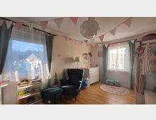 Helles Kinderzimmer mit zwei großen Fenstern, grauen Vorhängen und einer Wimpelgirlande an der Decke. Im Raum stehen ein Lesesessel mit Hocker, ein niedriges Bücherregal, eine weiße Kommode sowie Spielzeug und ein runder Teppich auf hellem Holzboden.