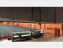 Blick von einer Bühne in einen leeren Vortragssaal mit mehreren Sitzreihen aus blauen Polstersesseln, Holzwänden und einem Rednerpult rechts. Auf der Bühne stehen Sofa-Sitzgruppen und ein Ständer mit Prospekten.