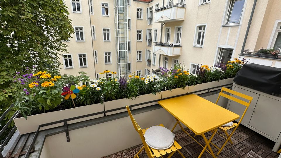 Blick von einem Balkon in einen Innenhof mit hellen Mehrfamilienhäusern, vielen Fenstern, kleinen Balkonen und einem gläsernen Aufzugschacht. Im Vordergrund stehen ein gelber Klapptisch mit zwei Stühlen auf Holzfliesen sowie Blumenkästen mit bunten Blumen entlang des Geländers.