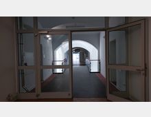 Ein heller Gebäudekorridor mit weißen Wänden und einem Fußboden aus flachen, dunklen Platten. Der Korridor führt durch eine Glastür zu einem erhöhten Gang mit roten Handläufen und einer hohen, weiß gewölbten Decke.