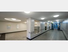 Ein moderner, heller U-Bahn-Bereich mit Fliesenwänden, grauem Bodenbelag und Deckenleuchten. Rechts befindet sich ein Aufzug mit Beschilderung, links sind Infotafeln an der Wand angebracht.