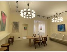 Heller Innenraum einer Galerie oder eines Ausstellungsraums mit cremefarbenen Wänden, modernen Kunstwerken an den Wänden und einem ovalen Holztisch mit Stühlen im Vordergrund. Zwei runde, moderne Kronleuchter mit vielen Glühbirnen hängen von der Decke, und im Hintergrund sind Regale sowie weitere Kunstwerke sichtbar.