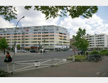 Straßenszene mit einem mehrstöckigen Wohn- und Geschäftsgebäude im Hintergrund, das Geschäfte im Erdgeschoss und Wohnungen in den oberen Etagen umfasst. Im Vordergrund sind eine Straße mit Fahrradständern, ein Gehweg sowie einige Bäume und Passanten zu sehen.