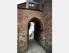 Backsteinbogen eines historischen Gebäudes mit gepflastertem Durchgang. Die Mauern bestehen aus ungleichmäßigem Mauerwerk aus Ziegeln und Steinen, während im Hintergrund Bäume und eine Straße mit Menschen sichtbar sind.