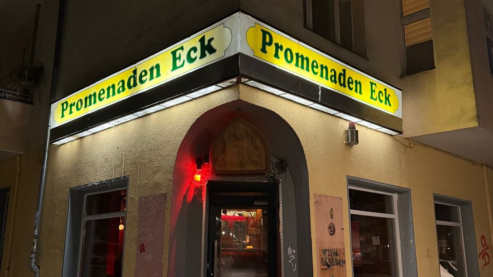 Eckeingang eines Mehrfamilienhauses bei Nacht mit beleuchtetem Eck-Schild „Promenaden Eck“ über einem Rundbogenportal und mehreren Fenstern. Vor dem Eingang liegt ein schmaler, verschneiter Gehweg; rotes Licht beleuchtet den Bereich um die Tür.