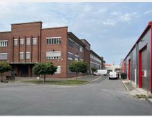 Zweigeschossiges Backsteingebäude mit Flachdach und vertikalen Fensterelementen links, angrenzend eine Straße mit geparkten weißen Transportern; rechts befinden sich moderne Lagerhallen mit roten Rahmen und Rolltoren. Im Hintergrund sind weitere Gebäude erkennbar.