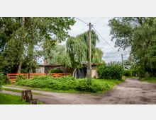 Ein kleines Gebäude mit Flachdach, umgeben von üppiger Vegetation und hohen Bäumen. Ein Weg aus Erde führt vorbei, daneben ein roter Holzzaun und ein Schild mit der Aufschrift „Zanderweg“.