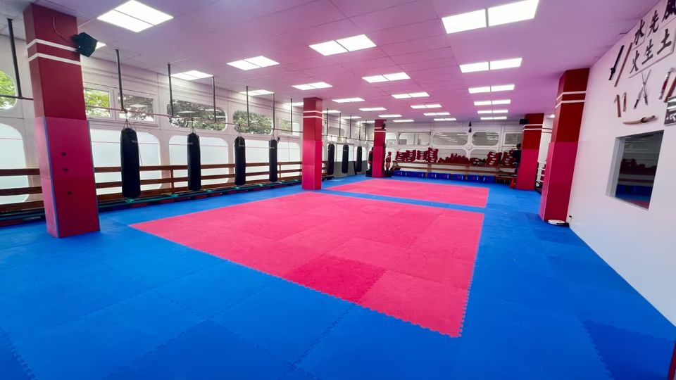 Ein großer Trainingsraum mit einer rot-blauen gepolsterten Bodenfläche, roten Säulen, an der Wand aufgehängten Boxsäcken und einer Spiegelwand. Der Raum hat eine Decke mit eingebauten Leuchtpaneelen und große Fenster mit Blick nach draußen.
