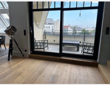 Blick aus einem modernen Wohnraum mit Holzfußboden durch eine bodentiefe Glas-Schiebetür auf eine Dachterrasse mit Fliesen, Sitzmöbeln und Geländer. Im Hintergrund ist eine städtische Gebäudekulisse unter blauem Himmel zu sehen.