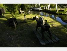 Rasenfläche mit historischen Werkzeugen und Geräten, darunter ein Schleifstein auf einem Holzgestell und eine alte Schubkarre, im Hintergrund ein kleiner Wasserkanal, gesäumt von Bäumen und umgeben von Grünflächen.