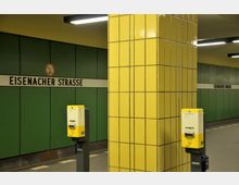 U-Bahn-Station mit grün gekachelten Wänden, einem weiß-schwarzen Stationsschild mit der Aufschrift "Eisenacher Straße" und gelben Säulen aus quadratischen Fliesen. Im Vordergrund stehen zwei gelbe Fahrkartenentwerter.