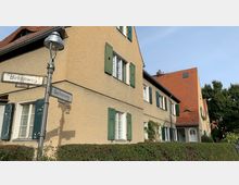 Straßenecke mit einem mehrstöckigen Wohngebäude aus beigem Putz und roten Ziegeldächern, Fenstern mit grünen Fensterläden und umliegender Hecke. Im Vordergrund steht eine Straßenlaterne mit Straßenschildern „Birkenweg“ und „Buchenweg“.