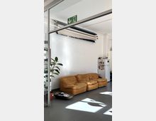 Innenraum eines Büros oder Studios mit weißer Wand, grauem Boden und einer Sitzgruppe aus drei niedrigen braunen Sofaelementen. Daneben stehen eine große Topfpflanze, ein kleiner Kühlschrank mit Magneten sowie über dem Sitzbereich mehrere aufgerollte Hintergrundrollen und ein grünes Notausgangsschild.