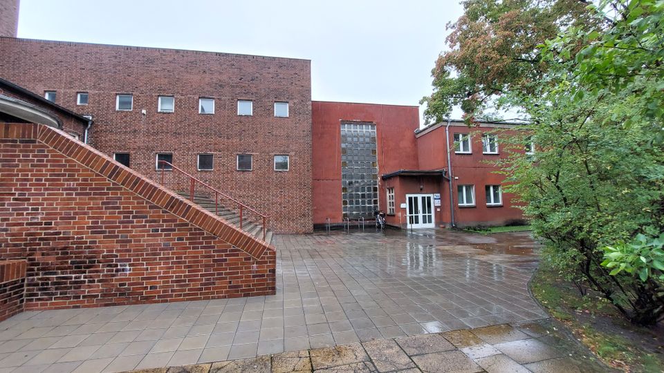 Backstein-Gebäudekomplex mit einer roten Fassade, rechteckigen Fenstern und einem großen Glasbaustein-Element in der Mitte. Davor ein gepflasterter, regennasser Hof mit Treppe und umgebenden Bäumen.