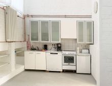 Kleine helle Küche in einem Raum mit weiß gestrichenen Ziegelwänden. Ausstattung umfasst weiße Schränke mit Glasfronten, einen Herd, eine Spüle, eine Kaffeemaschine, eine Spülmaschine und eine Kühl-Gefrier-Kombination; kupferne Rohrleitungen und ein Heizkörper an der Wand.