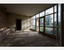 Ein leerer, verlassener Raum mit einer beschädigten, weiß gestrichenen Wand und freiliegenden Backsteinmauern auf der linken Seite. Die rechte Wand besteht aus großen Fenstern mit Metallgittern, durch die Sonnenlicht einfällt; eine Tür steht offen und führt zu einem Außenbereich mit Bäumen.