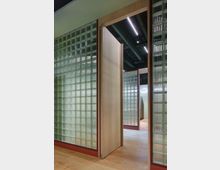 Innenraum mit Wänden aus Glasbausteinen in Metallrahmen, einer offenen Holztür und hellen Holzböden. Deckenstruktur mit sichtbaren Leitungen und integrierter Beleuchtung.