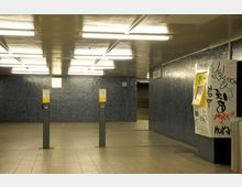 Unterführung mit blauen Fliesenwänden, erleuchtet durch rechteckige Deckenlampen. Im Vordergrund stehen zwei Entwerterautomaten, eine mit Graffiti übersäte Ticketmaschine ist rechts sichtbar, daneben eine Uhr an der Rückwand.