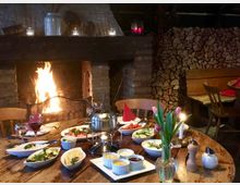 Gemütlicher Innenraum eines rustikalen Restaurants oder Hauses mit einem offenen Kamin, in dem ein Feuer brennt. Ein Holztisch ist mit Fondue-Set, Schalen mit Beilagen, Getränken und einer Vase mit Tulpen gedeckt; im Hintergrund ist gestapeltes Brennholz und eine Holzbank zu sehen.