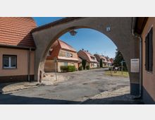 Straßenszene mit einer Häuserzeile aus beigefarbenem Putz und roten Ziegeldächern, durch einen großen, bogenförmigen Torbogen betrachtet. Die Umgebung wirkt dörflich mit gepflasterten Gehwegen und einer schmalen, leicht beschädigten Asphaltstraße.