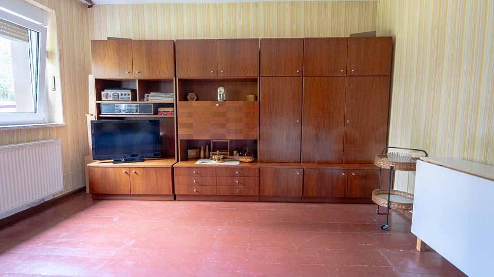 Innenraum eines Wohnzimmers mit großem, wandfüllendem Schrank aus dunklem Holz mit TV- und Regalfächern. Links ein Fenster über einem Heizkörper, gestreifte Tapete und roter Fliesenboden; rechts stehen ein kleiner Servierwagen und ein weißes Möbelstück.