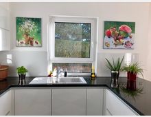 Moderne Küche mit weißem Unterschrank, einer schwarzen Arbeitsplatte und einem Edelstahlspülbecken unter einem Fenster, das auf einen Garten blickt. Zwei Gemälde mit Stillleben und grüne Topfpflanzen schmücken den Raum.