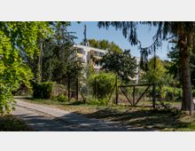 Gepflegter Gehweg neben einem Maschendrahtzaun, der ein von Bäumen und Sträuchern bewachsenes Grundstück umgibt, im Hintergrund ein mehrstöckiges Wohngebäude mit gelben und weißen Fassaden. Ein Mobilfunkmast steht auf dem Dach des Gebäudes.