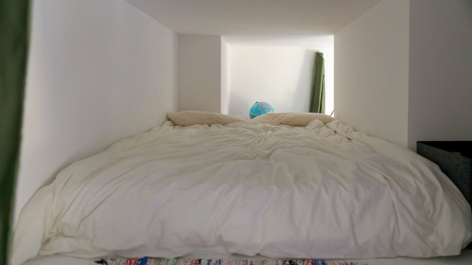 Schlafnische in einem minimalistischen Raum mit niedriger Decke und weißen Wänden, darin ein großes Bett mit weißer Bettwäsche. Am Fußende liegt ein gemusterter Teppich; hinten sind ein grüner Vorhang und ein blauer Globus zu sehen.