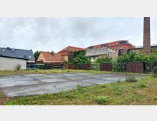 Leerer Asphaltplatz mit vereinzeltem Grasbewuchs, umgeben von niedrigen Metallzäunen und einer Mischung aus Wohnhäusern mit roten und grauen Dächern sowie einer hohen Backsteinschornsteinstruktur im Hintergrund.