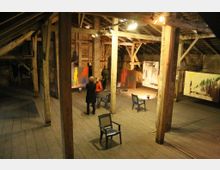 Ein geräumiger Dachboden mit Holzbalken und Holzboden, der als Ausstellungsraum genutzt wird. Auf den Wänden hängen abstrakte Gemälde, und im Raum stehen einzelne Stühle; wenige Personen betrachten die Kunst.