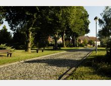 Ein gepflasterter Weg führt durch einen Park mit Bäumen, Bänken und Grasflächen. Im Hintergrund sind Häuser mit roten Ziegeldächern zu sehen, und eine Straßenlaterne steht am rechten Wegrand.