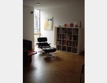 Wohnzimmer mit Holzfußboden, einer schwarzen Lounge-Chair-Sessel-Kombination, einem roten Couchtisch und einem Bücherregal an der rechten Wand. Ein großes Fenster mit Blick auf Bäume und ein Kunstwerk mit bunter Grafik hängen an der Wand.
