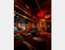 Innenraum einer Bar-Lounge mit dunklen Ledersofas und Sesseln um niedrige Tische auf einem gemusterten Teppich, neben großen Schaufenstern zur Straße. Kassettierte Decke mit ornamentalen Feldern und warmem, rötlichem Licht sowie Holzrahmen und Glastür im Hintergrund.