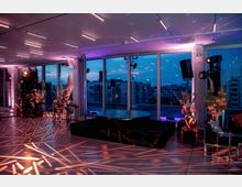 Moderner Veranstaltungsraum mit großen Glasfenstern, die den Blick auf eine städtische Skyline bei Abenddämmerung freigeben. Im Vordergrund befinden sich eine runde Bühne, dekorative Blumenarrangements und Beleuchtungselemente, die Muster auf den Boden projizieren.