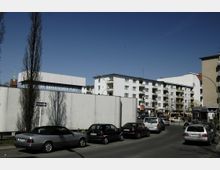 Straßenszene mit parkenden Autos und mehrstöckigen Wohngebäuden im Hintergrund. Auf einer weißen Wand im Vordergrund steht „Bayerischer Platz“, und ein kleiner Teil eines Kirchturms ist links sichtbar.