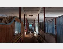 Innenraum eines Stalls mit Holz- und Metallboxen auf beiden Seiten eines zentralen Ganges. Die Decke ist aus Holzlatten, rote Pfeiler stützen den Raum, und links liegt gestapeltes Stroh.