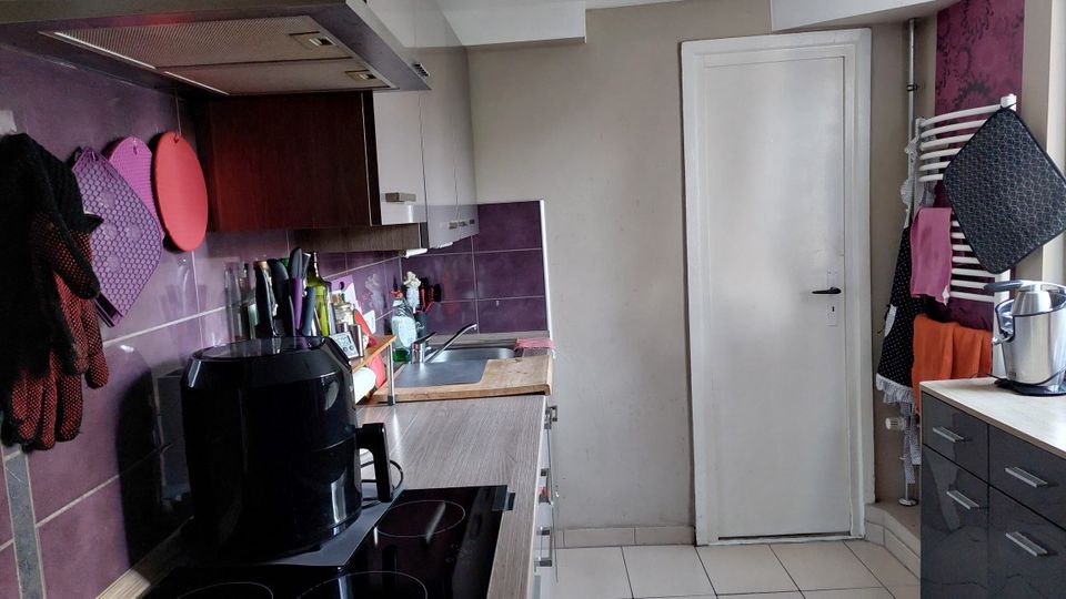 Schmale Küche mit L-förmiger Arbeitsfläche, Herd mit Dunstabzug und Spüle, flankiert von weißen und dunklen Schränken. Violette Fliesenrückwand und Wandpaneele, helle Bodenfliesen sowie eine weiße Tür am Ende des Raums; an der rechten Wand hängen Handtücher und Küchenutensilien.