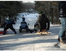 Eine winterliche Rodelbahn in einem schneebedeckten Waldgebiet, mit mehreren Personen, die auf Schlitten sitzen oder stehen und sich auf die Abfahrt vorbereiten; im Hintergrund sind weitere Menschen und ein kleines Gebäude sichtbar.