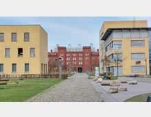 Backsteingebäude mit historischer Architektur im Hintergrund, eingerahmt von zwei modernen, gelbbraunen Bürogebäuden. Im Vordergrund befinden sich ein gepflasterter Weg, Grünflächen mit Sitzbänken und verteilt liegende Steine auf einem gepflasterten Platz.