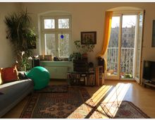 Wohnzimmer mit Holzfußboden, bunt gemusterten Teppichen, einer Couch mit Kissen, großen Zimmerpflanzen und einem grünen Gymnastikball. Fenster und eine Balkontür mit gelbem Vorhang lassen Tageslicht herein und bieten Ausblick auf Bäume und einen Balkon mit Pflanzen.